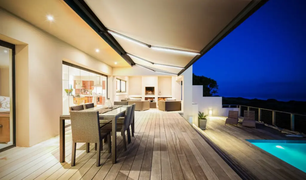 Luxury outdoor dining space with a modern retractable awning, warm lighting, and a poolside view during evening time.