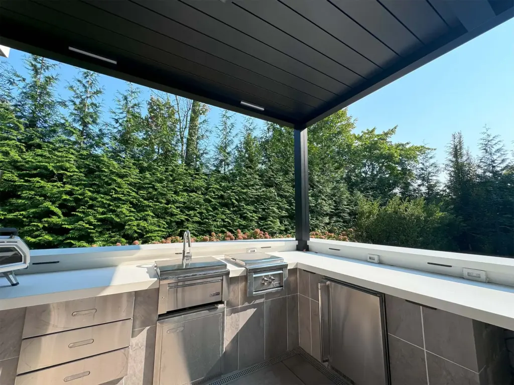 A modern pergola providing shade over an outdoor kitchen with built-in counters and a sink, surrounded by tall green trees.