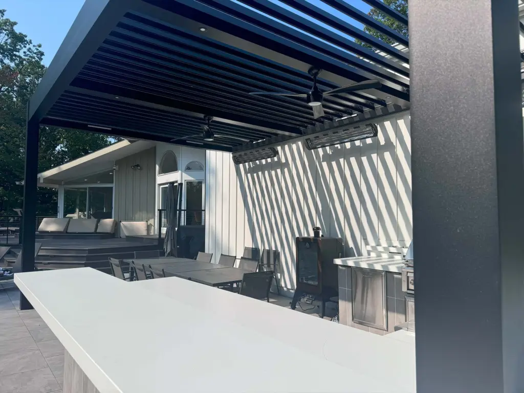 A sleek black louvered pergola shading an outdoor kitchen and bar area, with sunlight casting clean line patterns across the countertop and patio.