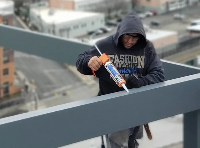 Worker applying sealant to a steel beam on a rooftop construction site with buildings visible in the background.