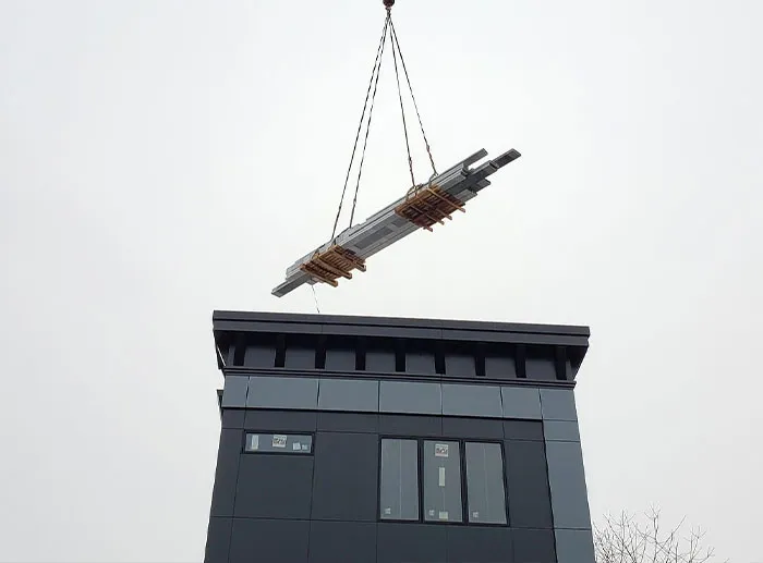 A crane lifting a large louvered roof frame above a modern building during installation.