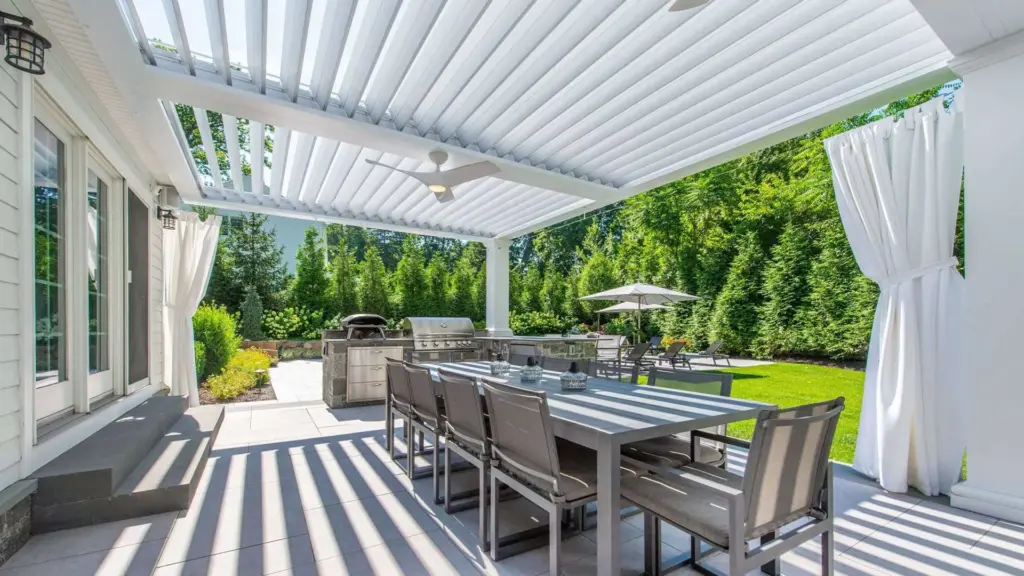Outdoor dining space beneath a white louvered pergola with patio furniture, ceiling fan, and a backyard garden view.