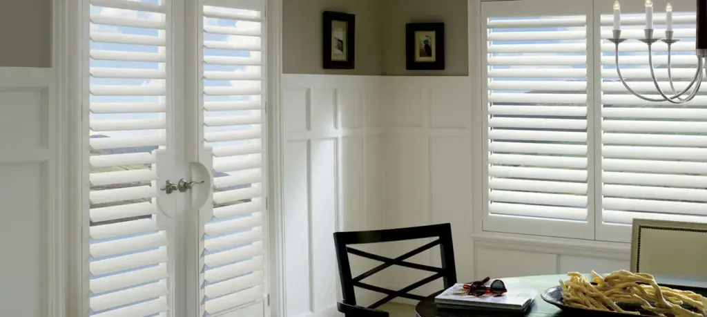 Bright dining room featuring white plantation shutters on windows and a glass door, allowing soft natural light to enter.