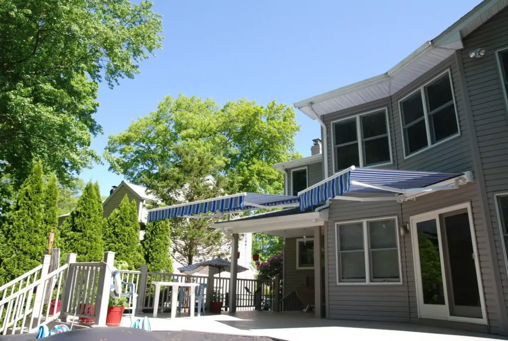 Blue retractable awning extended over the patio area of a gray two-story home surrounded by trees and outdoor seating.