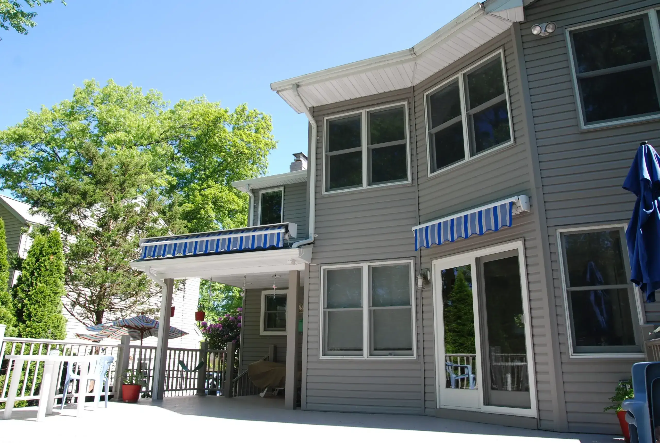 Blue-striped retractable awning extended over the patio of a gray two-story home surrounded by greenery.