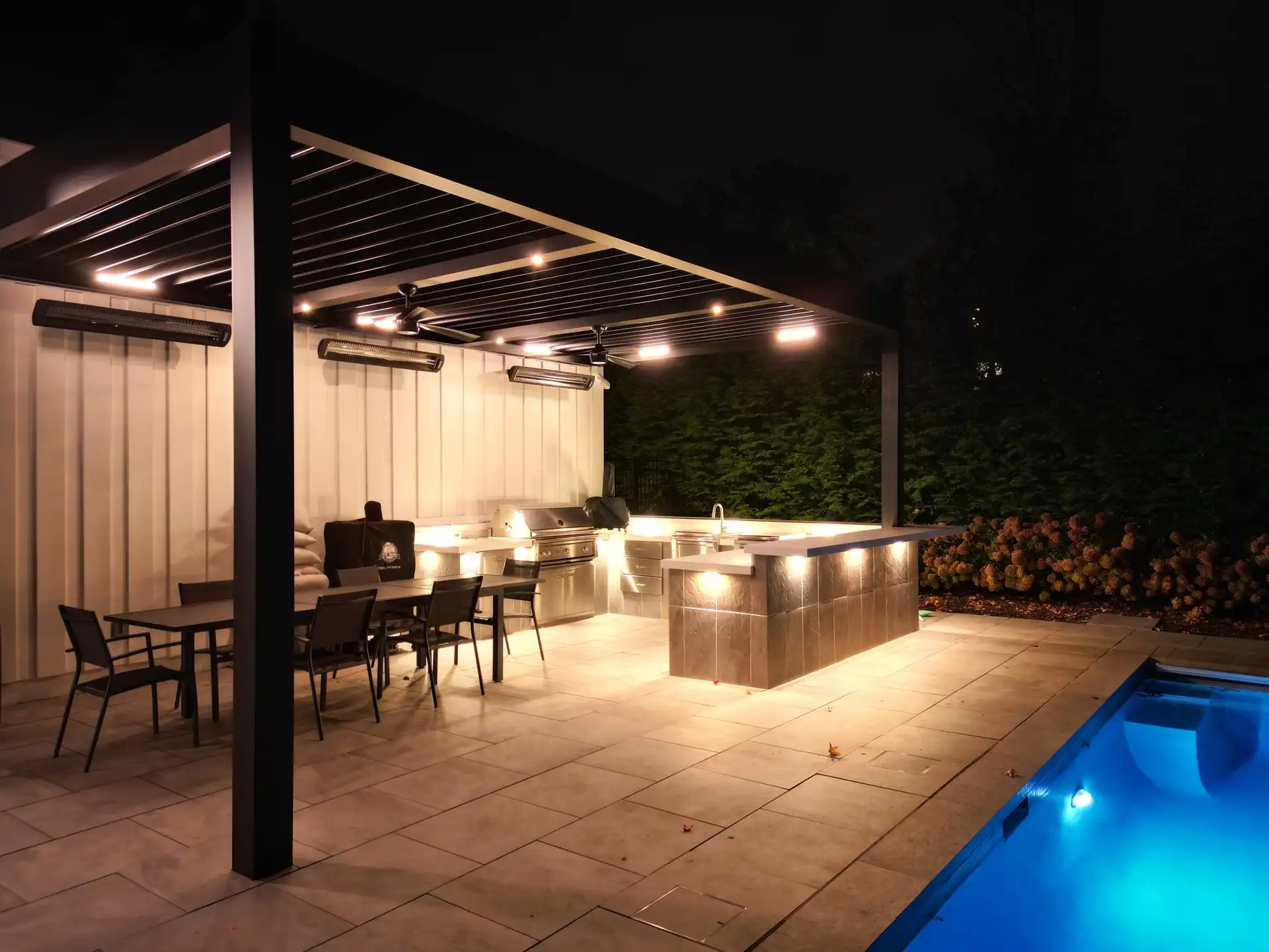 A modern outdoor pergola illuminated at night with built-in lights, featuring a dining area and bar beside a glowing pool.